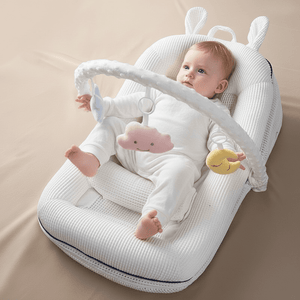 Baby lying on a white textured cushion with toys, sitting up with support from a white crib safety bar.