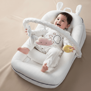 Baby sitting in a white baby bouncer with toys on a beige blanket