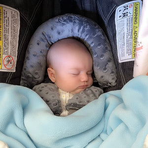 A newborn baby sleeping in a car seat, equipped with a white and blue sleeping pillow with a soft plush toy attached to the baby's stroller.