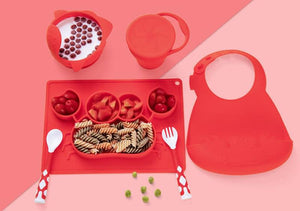 Red children's mealtime set including a tray, bowl, cup, bib, and utensils on a pink background.