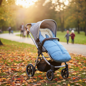A stroller with a light blue quilted footmuff and gray canopy sits on a grassy path covered in fallen autumn leaves, with blurred people walking in the background during golden hour.