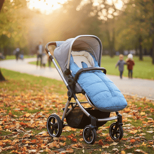 A stroller with a light blue quilted footmuff and gray canopy sits on a grassy path covered in fallen autumn leaves, with blurred people walking in the background during golden hour.