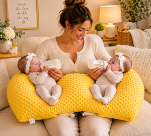 Woman holding two babies with bottles on a yellow pillow in a cozy living room.