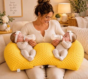 Woman holding two babies with bottles on a yellow pillow in a cozy living room.