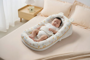 A baby bassinet with a soft, plush padded border featuring a light beige, white, and pastel autumn-themed animal print, shown on a bed with a wooden bedside table in the background.