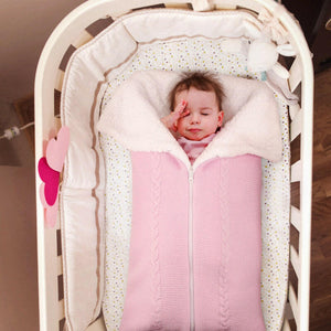 A baby sleeping in a crib, covered with a pink and white knitted sleeping bag blanket.