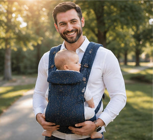 A man wearing a white long-sleeve shirt carries a baby in a navy blue baby carrier with a subtle gold speckled pattern, set against a blurred park background.