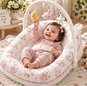A baby sits in a padded bassinet-style baby chair with floral fabric, holding a yellow moon toy under a white arch. The room is softly lit with a beige rug, flowers, and toys in the background.