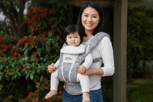 A man wearing a white shirt carries a baby in a gray and white star-patterned baby carrier outdoors. The baby is dressed in a white long-sleeve top and pants.