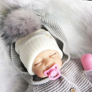 A baby wearing a gray knitted winter hat with a faux fur brush on a white furry background.