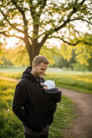 A man wearing a black baby carrier hoodie with a baby inside, standing on a park path with a large tree in the background during golden hour.