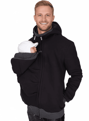 A smiling man wearing a black hooded jacket with a gray inner hood and pocket, holding a baby wrapped in a white blanket against his chest, set against a plain white background.