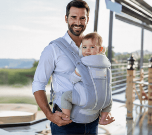A man wearing a white shirt and blue jeans carries a baby in a light gray and white multi-position baby carrier. The photo is taken outdoors on a balcony with a railing and a blurred background.