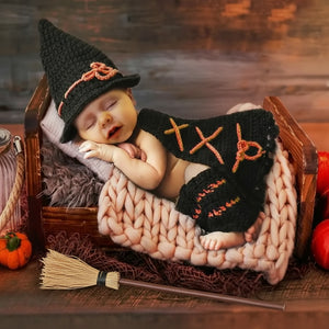 Newborn baby in Halloween-themed outfit with witch hat and scarf, lying in a wooden crate.