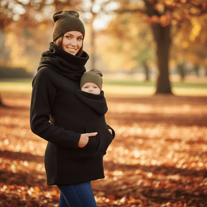A woman wearing a black hooded winter coat with a built-in baby carrier and matching black cowl neck warmer, carrying a baby in an outdoor autumn park with fallen leaves.