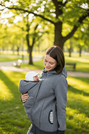 A woman in a gray baby carrier hoodie holds a baby in a white hat in a sunny park with green trees.