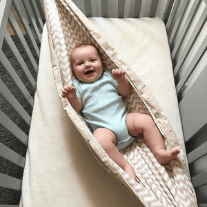 Baby in a light blue onesie lying on a patterned mat inside a crib.