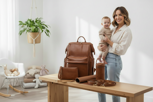 Woman holding a baby next to a set of brown leather bags and accessories on a wooden table.