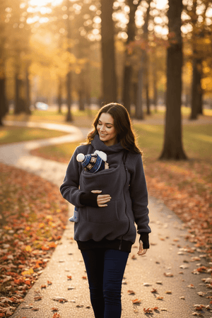 A woman walking on a leaf-strewn park path wearing a dark gray baby carrier hoodie and carrying a baby in a baby carrier.