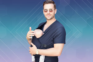 Person with tattoos holding a baby in a sling against a white background