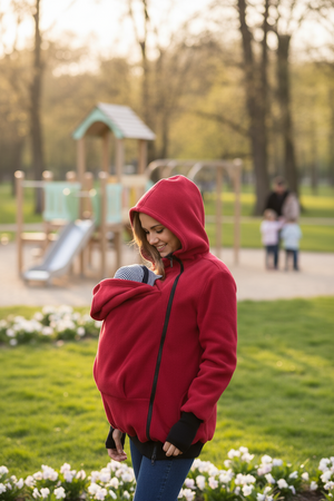 Woman in a red hoodie carrying a baby in a park with playground equipment in the background