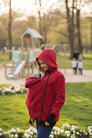 Woman in a red hoodie carrying a baby in a park with playground equipment in the background