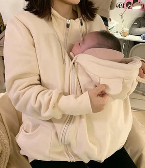A person wearing a beige kangaroo-style baby carrier hoodie with a baby inside.