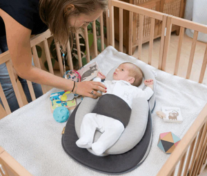 A newborn baby lying in an anti-reflux elevated baby lounger with a woman attending to the baby. The lounger is placed in a crib with toys around and has a gray color scheme.