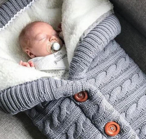 A baby wrapped in a gray sleeping bag with a cable knit pattern and button closures, lying on a couch.