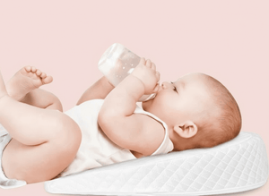 Baby lying on a white pillow, holding a bottle against a pink background