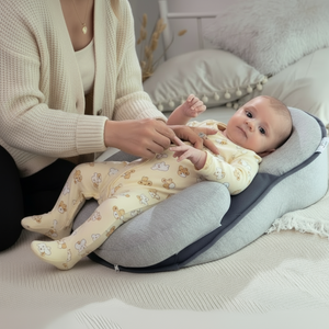 Baby lying on a gray and blue baby lounger pillow with a person's hand gently touching the baby's foot, all on a light-colored couch.