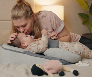 Woman lying on a bed with a baby lounger surrounded by toys, in a cozy bedroom setting.