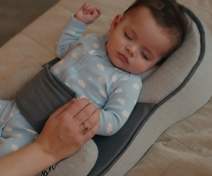 Baby in a blue onesie with white polka dots lying on a gray baby lounger, held by an adult hand.