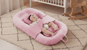 Baby in a pink bassinet in a nursery with a crib and toys in the background.