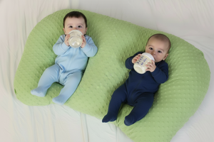 Two babies in pink outfits lying on a blue pillow, each holding a bottle.