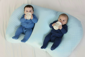 Two babies in pink outfits lying on a blue pillow, each holding a bottle.