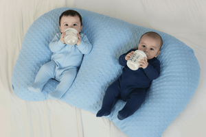 Two babies lying on a blue baby pillow, each holding a bottle.