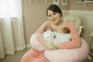 Woman holding two newborn babies wrapped in white blankets in a room with light blue walls and framed pictures.
