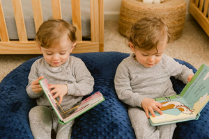 Two children sitting on a blue cushion, each reading a book in a nursery setting.