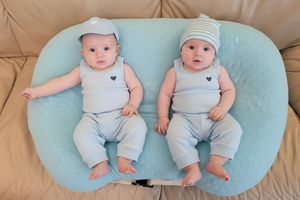 Two babies in matching outfits sitting on a blue cushion on a beige couch.