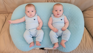 Two babies in matching outfits sitting on a blue cushion on a beige couch.