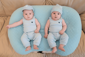Two babies in matching outfits sitting on a blue cushion on a beige couch.