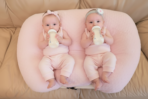 Two babies in pink outfits sitting on a blue cushion with a beige couch background