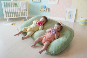 Two babies lying on a green cushion with toys around them.