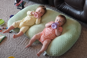 Two babies lying on a green pillow with toys around them.