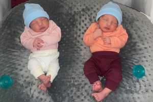 Two babies wearing hats and outfits on a textured surface with toys.