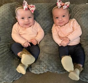 Two babies wearing matching outfits and headbands sitting on a cushion.