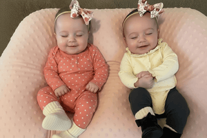 Two babies wearing matching outfits and headbands sitting on a cushion.