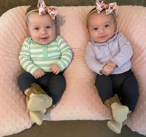 Two babies sitting on a pink cushion wearing headbands with floral designs.