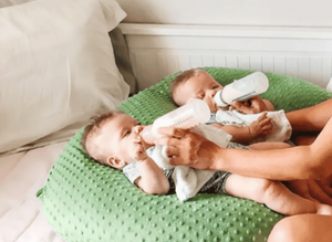Two babies lying on a cushion with a person holding bottles